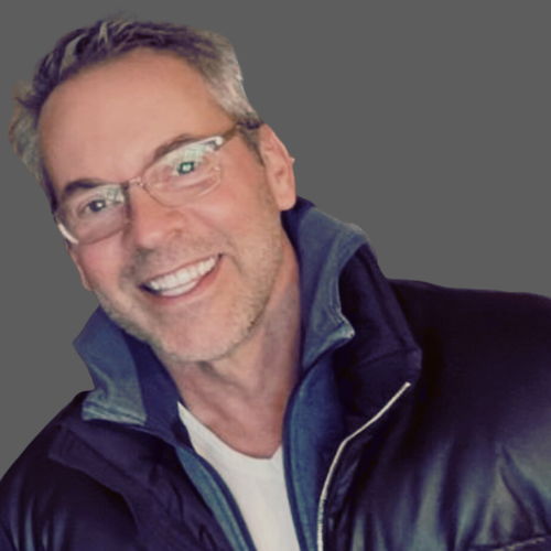 Mark smiling, wearing glasses and a dark jacket, against a plain gray background.